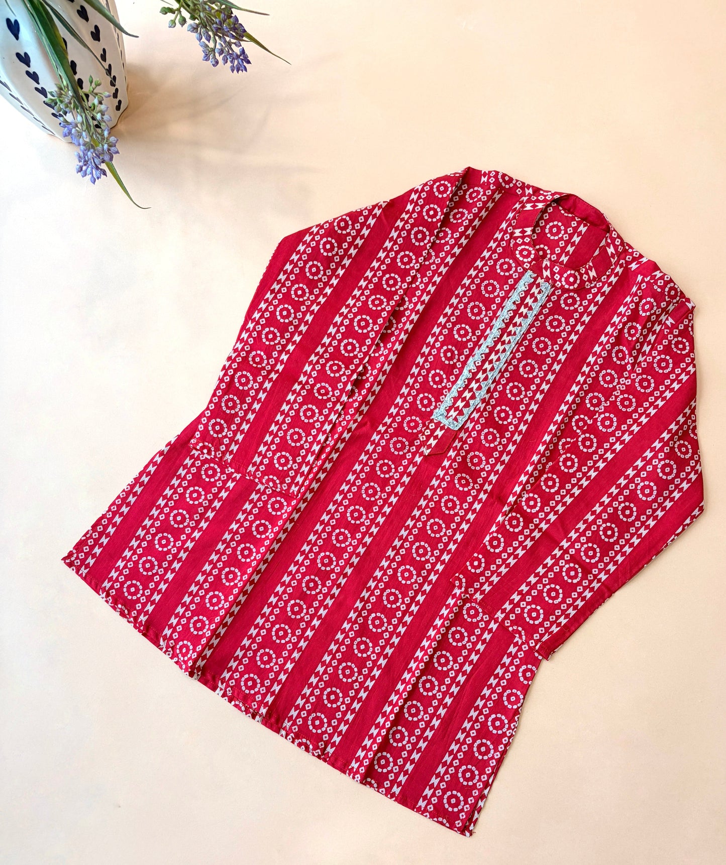 A red long-sleeve kurta with vertical white and pink geometric patterns, laid flat on a light beige background with a small potted plant in the top-left corner.