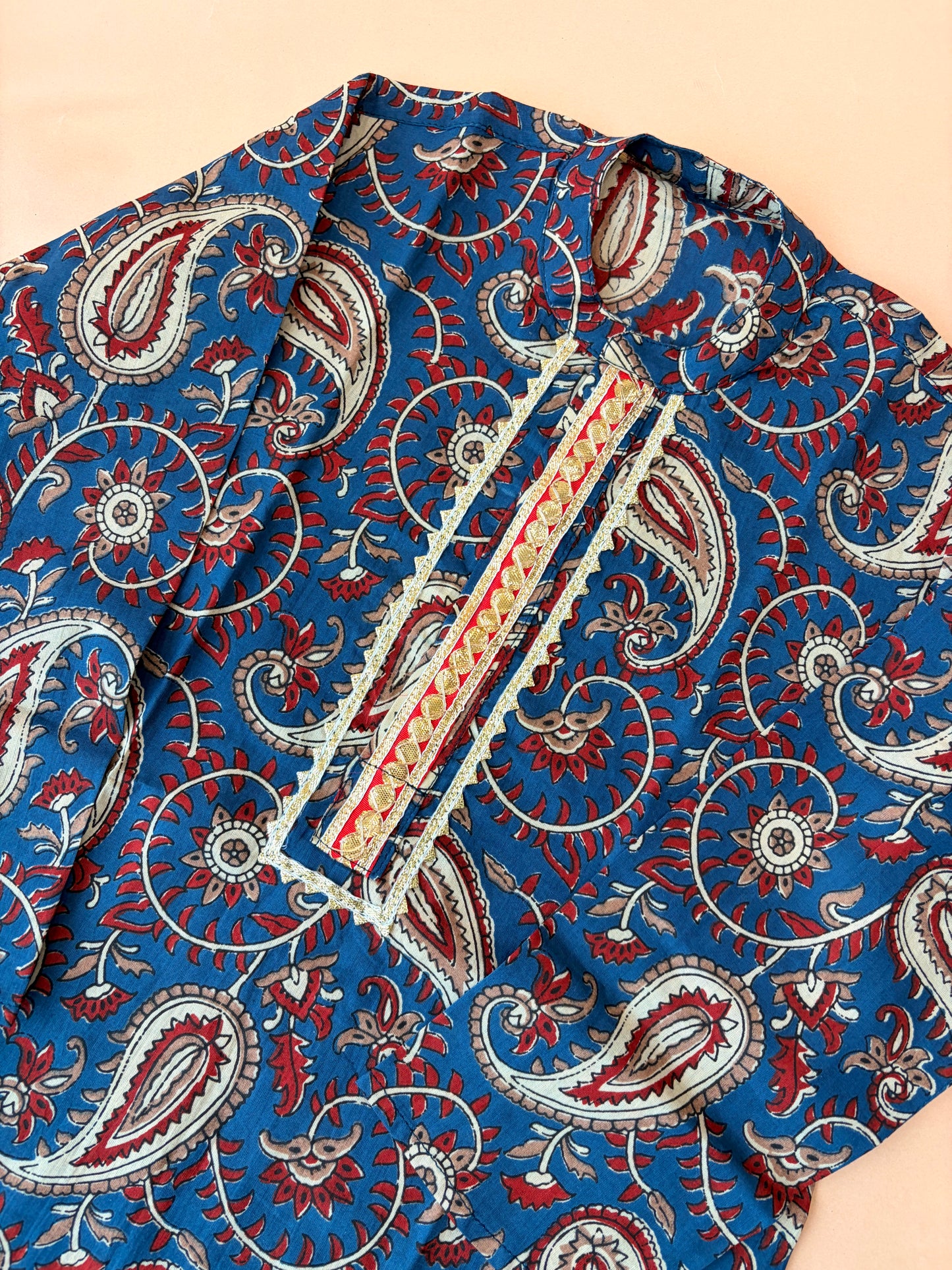 A close-up of a blue fabric garment with an intricate red, white, and beige paisley block print pattern and a decorative embroidered placket.
