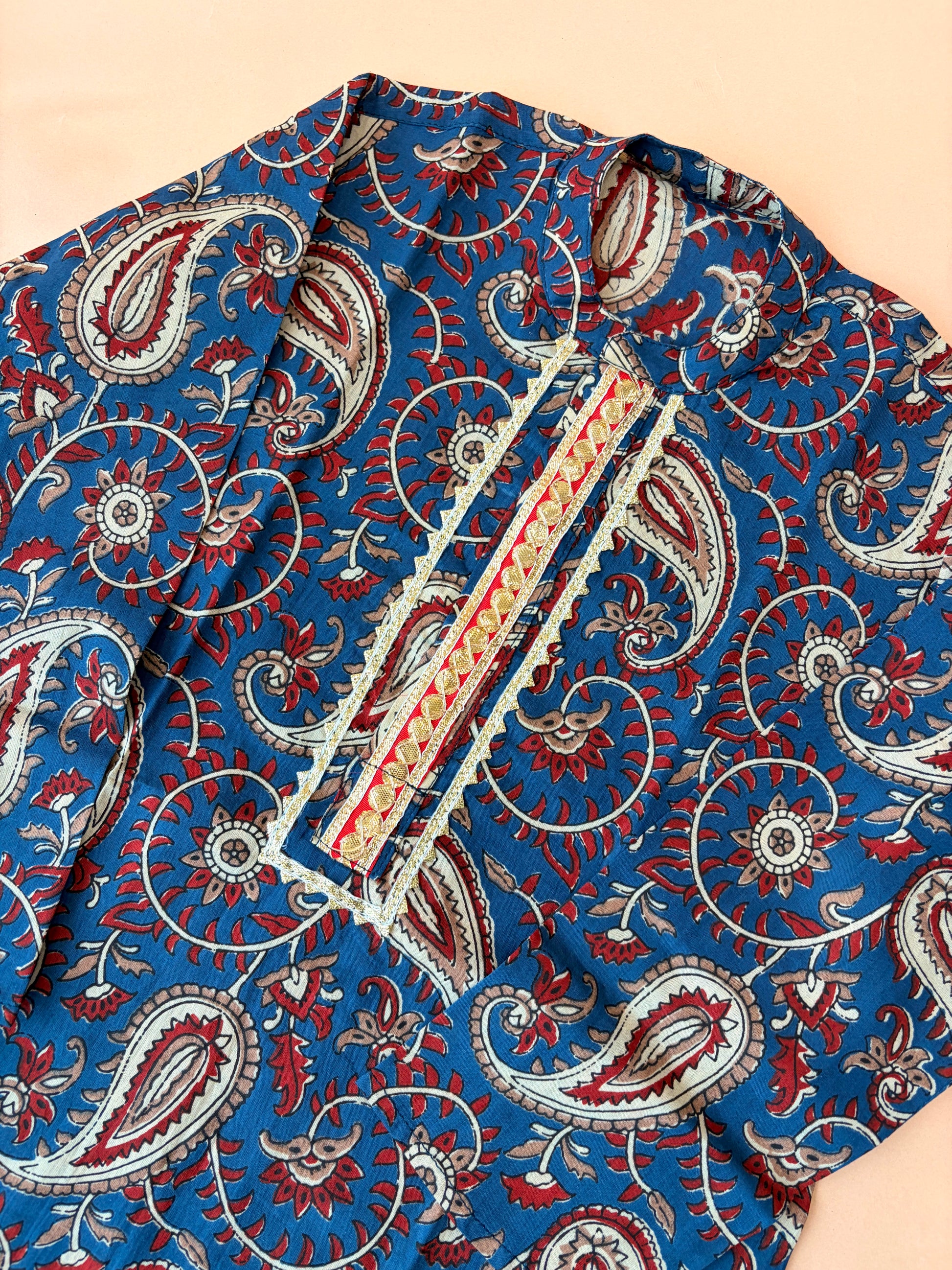 A close-up of a blue fabric garment with an intricate red, white, and beige paisley block print pattern and a decorative embroidered placket.