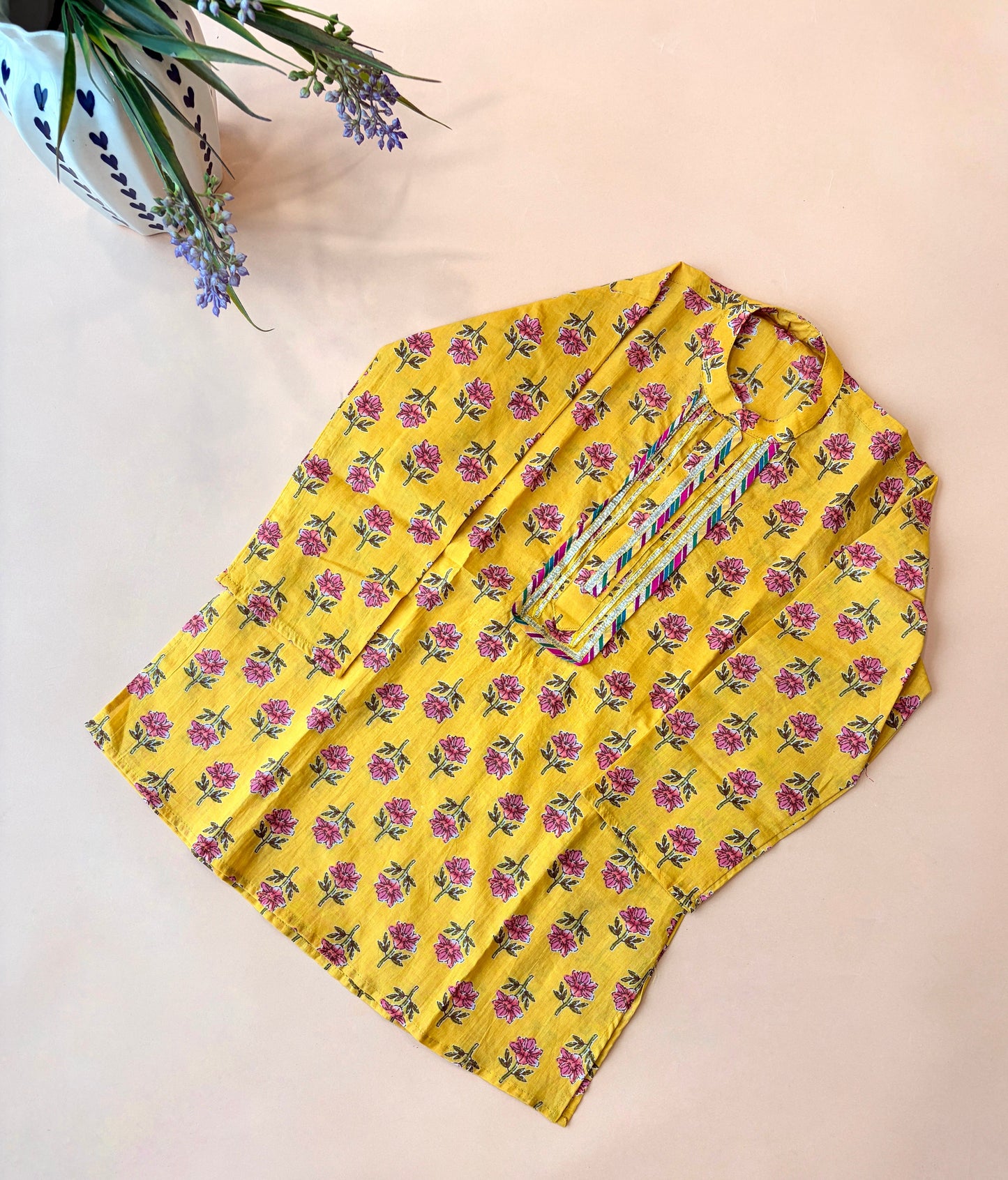 A yellow kurta with a pink and green floral print, laid flat on a light beige background with a decorative plant in the top-left corner.