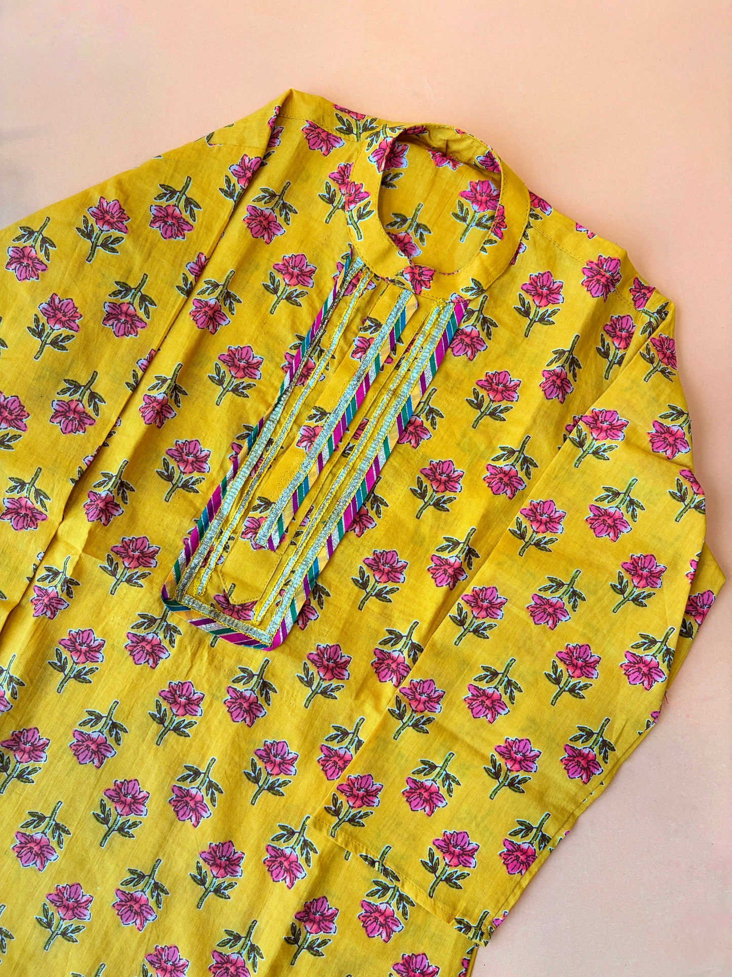 A yellow kurta with an all-over pink and green floral print, featuring a multicolored embroidered placket on the chest, laid flat on a light peach background.