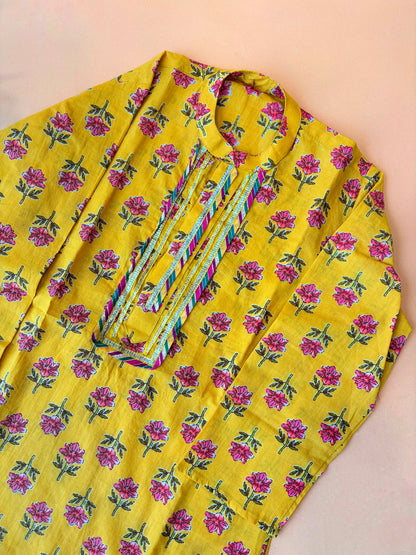 A yellow kurta with an all-over pink and green floral print, featuring a multicolored embroidered placket on the chest, laid flat on a light peach background.